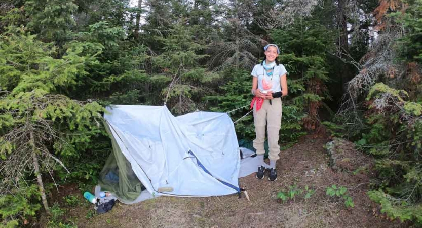 an outward bound student stands beside their tarp shelter in a wooded area 